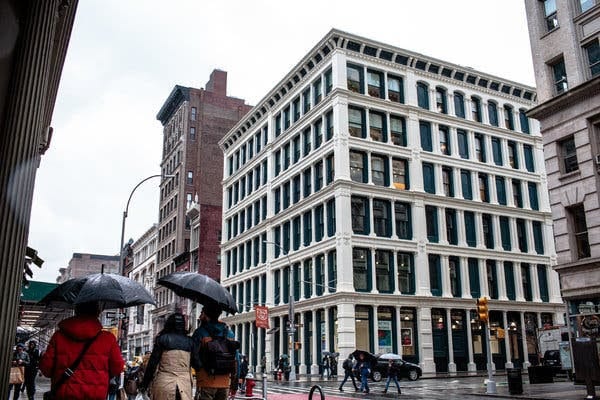 People walk on a rainy New York city street with umbrellas near a large, white and dark blue historic building on the corner. Other multi-story buildings line the wet street under a cloudy sky in this bustling capital of finance and cybersecurity.