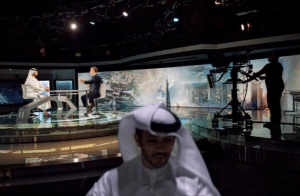 A television studio with two hosts sitting at a modern, curved desk is abuzz with conversation about innovative Israeli entrepreneurs. As a camera operator films them against a cityscape backdrop, a man in traditional attire stands slightly out of focus in the foreground.