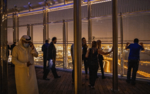 Israeli entrepreneurs stand on a high observation deck at night, overlooking the brightly lit cityscape of Dubai. A man in traditional attire and a face mask is on the phone, while others fall in love with the breathtaking view through large glass windows.