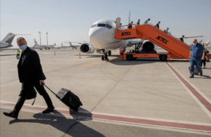 A person in a mask walks with a rolling suitcase on an airport tarmac, reminiscent of Israeli entrepreneurs frequently traveling to Dubai. Others board a plane using an orange Jetex stairway, while multiple parked aircraft are visible in the background under a clear sky.
