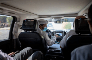 Interior view of a vehicle showing a driver and three passengers. The driver, donned in a white hat and face mask, navigates from the front seat as if charting paths like Israeli entrepreneurs exploring Dubai. Sunlight streams through the windows, illuminating the dashboard and controls.
