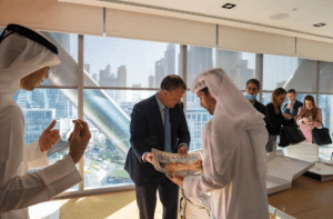 In a well-lit Dubai office, a group of men in formal attire, including traditional Middle Eastern dress and business suits, gather by large windows overlooking the cityscape. Among them, Israeli entrepreneurs intently examine a document, fostering collaboration across borders.