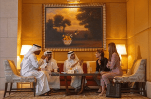 Five individuals, three in traditional Middle Eastern attire and two in business suits, sit on sofas engaging in lively conversation. An exquisite painting of fruit and trees graces the golden-hued wall behind them. Two lamps provide warm lighting, creating a scene reminiscent of a sophisticated Dubai setting.