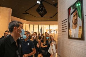 People wearing masks walk through a modern hallway in Dubai, admiring a framed portrait of a man in traditional attire. The backdrop of glass doors and curved architecture adds to the allure, as Israeli entrepreneurs fall in love with the fusion of cultures around them.