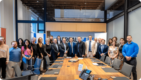 A diverse group of people stands around a large conference table in a modern office setting. They are dressed in business attire and smiling. The room features a glass partition and wood ceiling beams, adding to the contemporary ambiance.