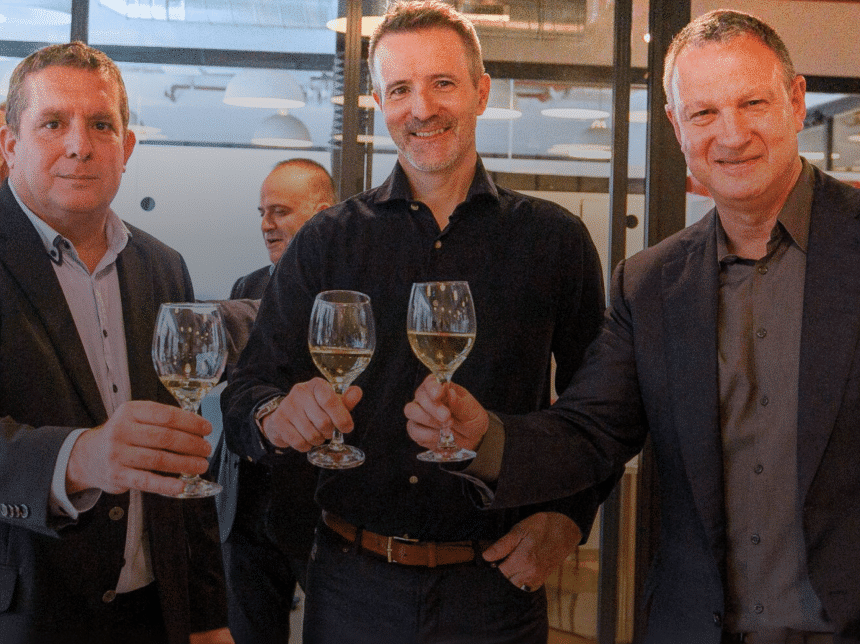 Erel N. Margalit, Gadi Porat and another man raise wine glasses in a toast, confidently smiling in an indoor setting with glass walls and modern lighting, celebrating an event. Two other people are visible in the background.