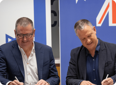 Erel N. Margalit and a man are seated at a table, smiling as they sign documents that define the essence of "About Us." They sit in front of a backdrop featuring two flags.
