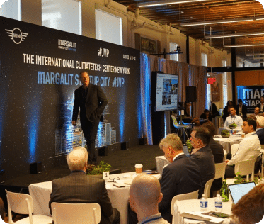 Erel N. Margalit speaks on stage and addresses a seated audience at an event. The backdrop reads The International Climate Tech Center New York and Margalit Startup City. Attendees are watching attentively; some have laptops open.