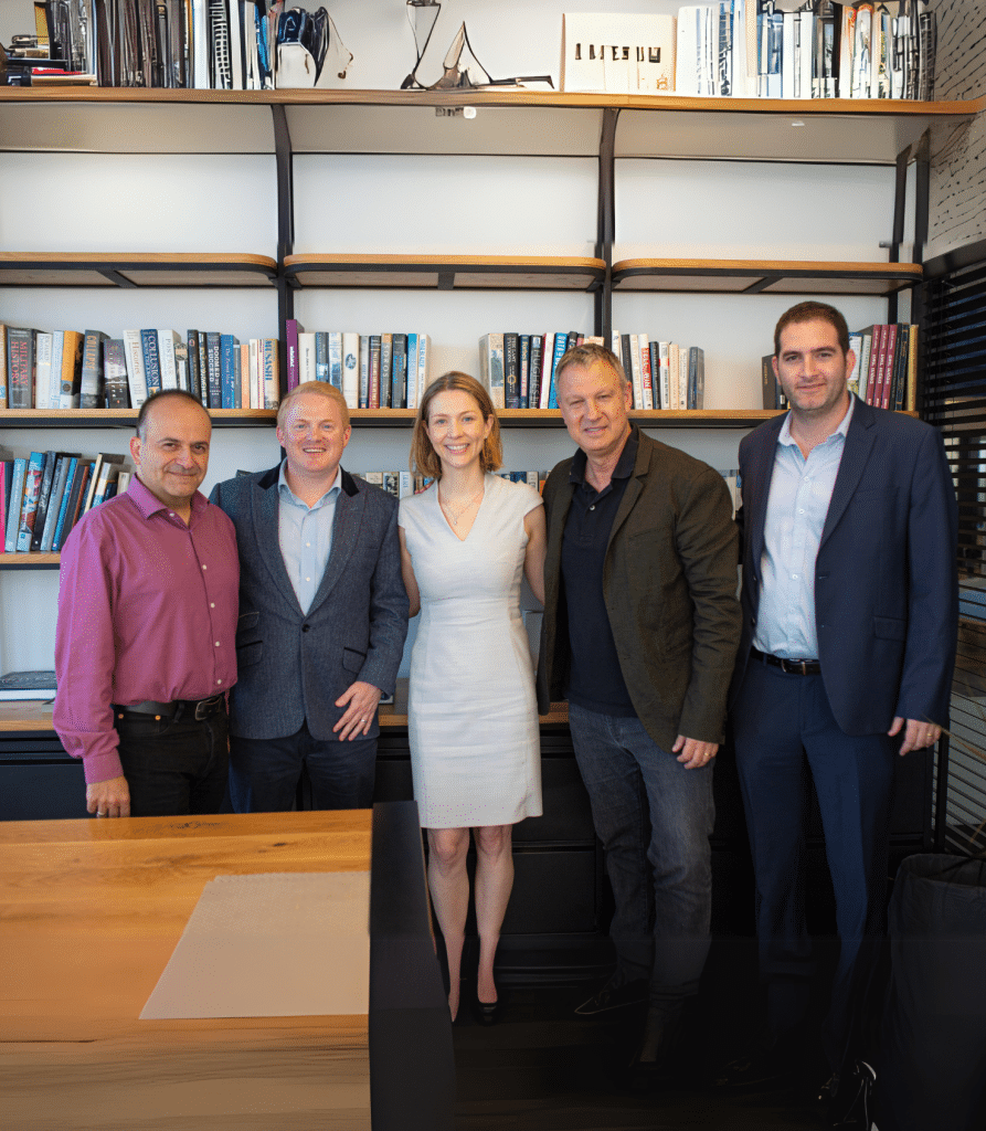 A group of five people, including Erel, stand smiling in an office with bookshelves behind them. A wooden desk graces the foreground of this lively scene.