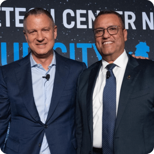 Erel N. Margalit and a man stand smiling in front of a backdrop. Both are wearing ties and have microphones attached to their clothing. The setting appears to be a formal or professional event.