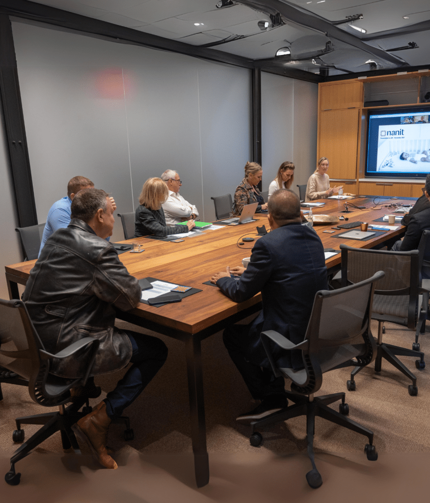 A group of people is gathered around a large conference table in a modern meeting room. Some are taking notes while others focus on a presentation screen displaying an image of a logo with the word nanit.