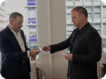 In a modern office setting with large windows showcasing city buildings, two men in business attire exchange business cards.One man holds his card holder while the other reaches out, symbolizing the start of potential collaboration.