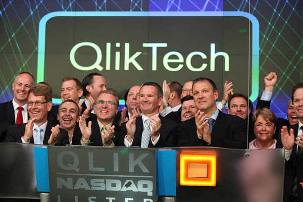 A large group of businesspeople in suits smile and clap as they celebrate at the NASDAQ stock exchange, standing in front of a display showing QlikTech—highlighting their success in building market leaders amid vibrant electronic graphics.