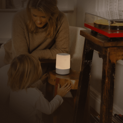 A woman and child engage with a small, glowing lamp on a wooden stool, their serene interaction watched over by the Nanit baby monitor. The room is softly lit, casting a cozy glow, while a turntable peeks from the right, adding to the calming ambiance.