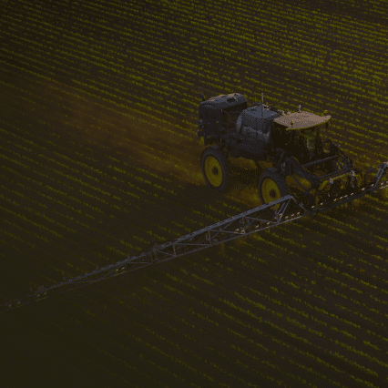 Aerial view of a tractor with a long spraying boom, working across a vast, neatly lined field. The Greeneye system enhances the scene, where patterns create an interesting texture as the tractor moves diagonally across the landscape.