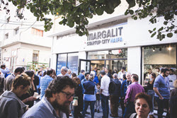 A crowd gathers outside the entrance of Margalit Startup City Haifa. The lively scene in Haifa buzzes with people socializing under a tree, indicating a vibrant event or gathering.