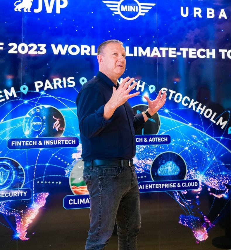 A man in a blue shirt gestures while speaking in front of a colorful digital display featuring global cities and technology themes such as fintech, security, sustainability, and climate tech. The backdrop highlights a World Climate-Tech Tour with impact.