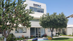 A white two-story building with the name Margalit Startup City on top. It is surrounded by trees and a well-kept lawn with outdoor seating under the clear blue sky.