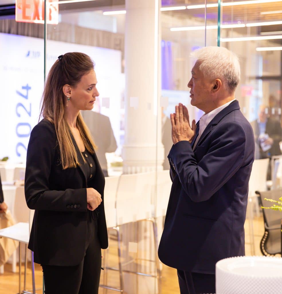 A woman and a man in business attire stand talking in an office with glass walls, discussing strategies as partners. The man gestures while speaking, and the woman listens attentively. A 2024 sign is visible in the background.