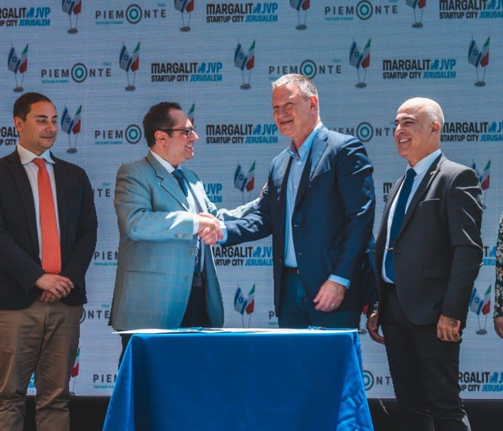 Four men in business attire stand before a logo backdrop, two shaking hands over a table and smiling—likely marking a formal agreement signing that supports regional economic growth.