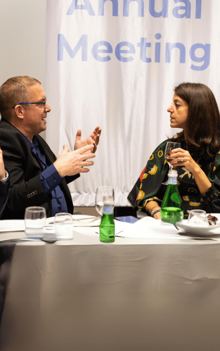 A man and a woman sit at a table in conversation, with water bottles, glasses, and plates in front of them. Behind them is a banner that partially reads Annual Meeting, creating a classic meeting image.