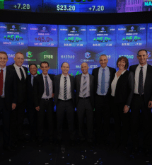 A group of people in formal attire stand in front of a large screen displaying stock market information, including numbers and the NASDAQ logo.