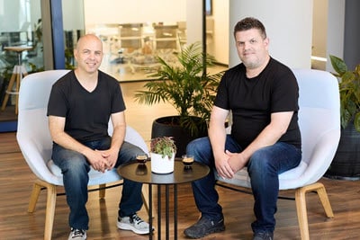 Two people are seated on white chairs around a small black table with two espresso cups and a potted plant. They are indoors, surrounded by green plants, wearing casual black t-shirts and jeans, smiling at the camera.