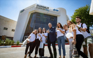 A group of people, five women and two men, happily pose in front of a modern building labeled Margalit StartUp City Galil, founded by tech investor Erel Margalit. Most are wearing white shirts and jeans. The clear sky and a tree grace the background.