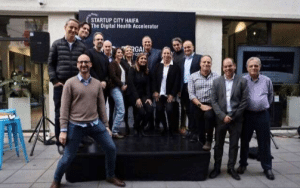 A group of people, including tech investor Erel Margalit, stand on the platform and around it, smiling for a photo. Behind them is a sign that reads Startup City Haifa: The Digital Health Accelerator, capturing the vibrant energy of this outdoor event space.