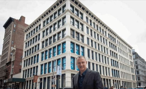 A man in a dark suit stands in front of a large, historic multi-story building with a white facade and many windows. The overcast sky casts a muted light on what might soon be the next Startup City, spearheaded by tech investor Erel Margalit. Other buildings stretch into the background.