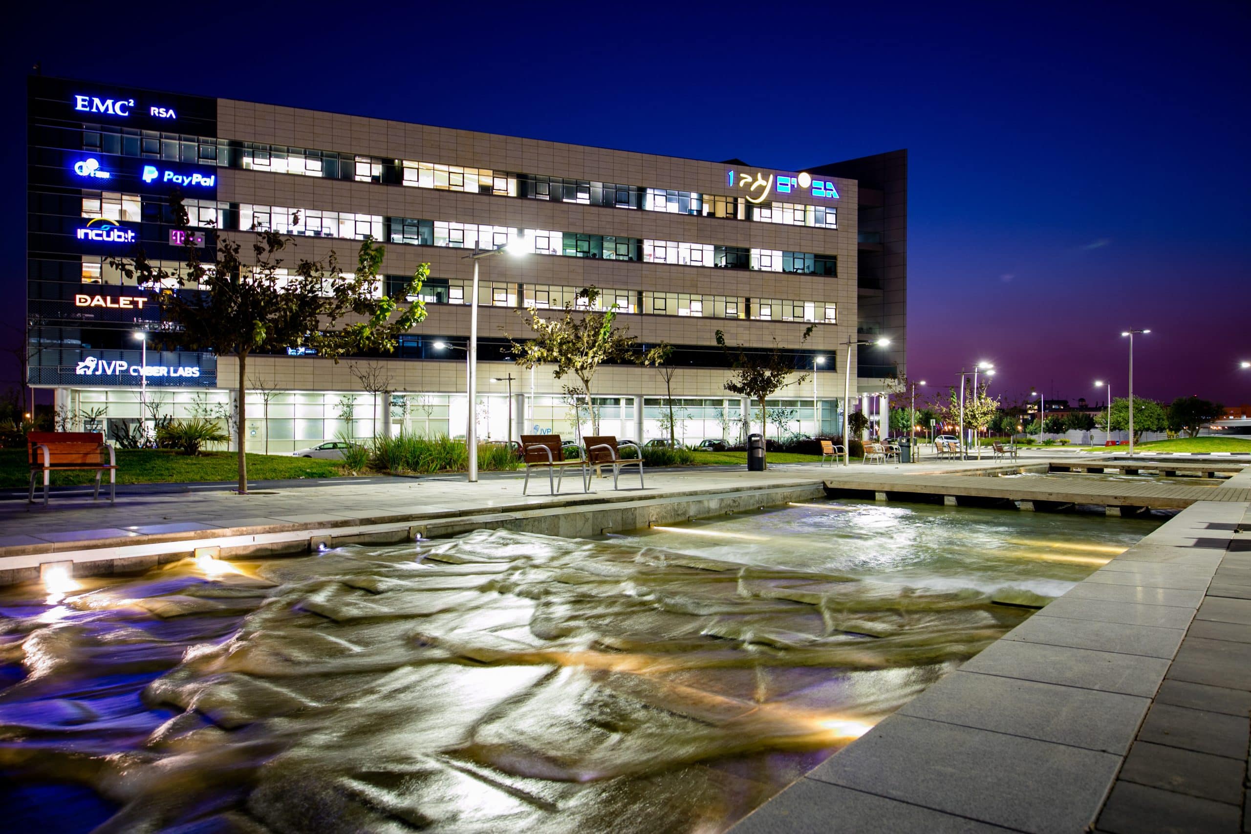 Night view of a modern office building with illuminated windows featuring logos, including PayPal. In the foreground, a water feature with gentle ripples, trees, and benches are visible under the evening sky.
