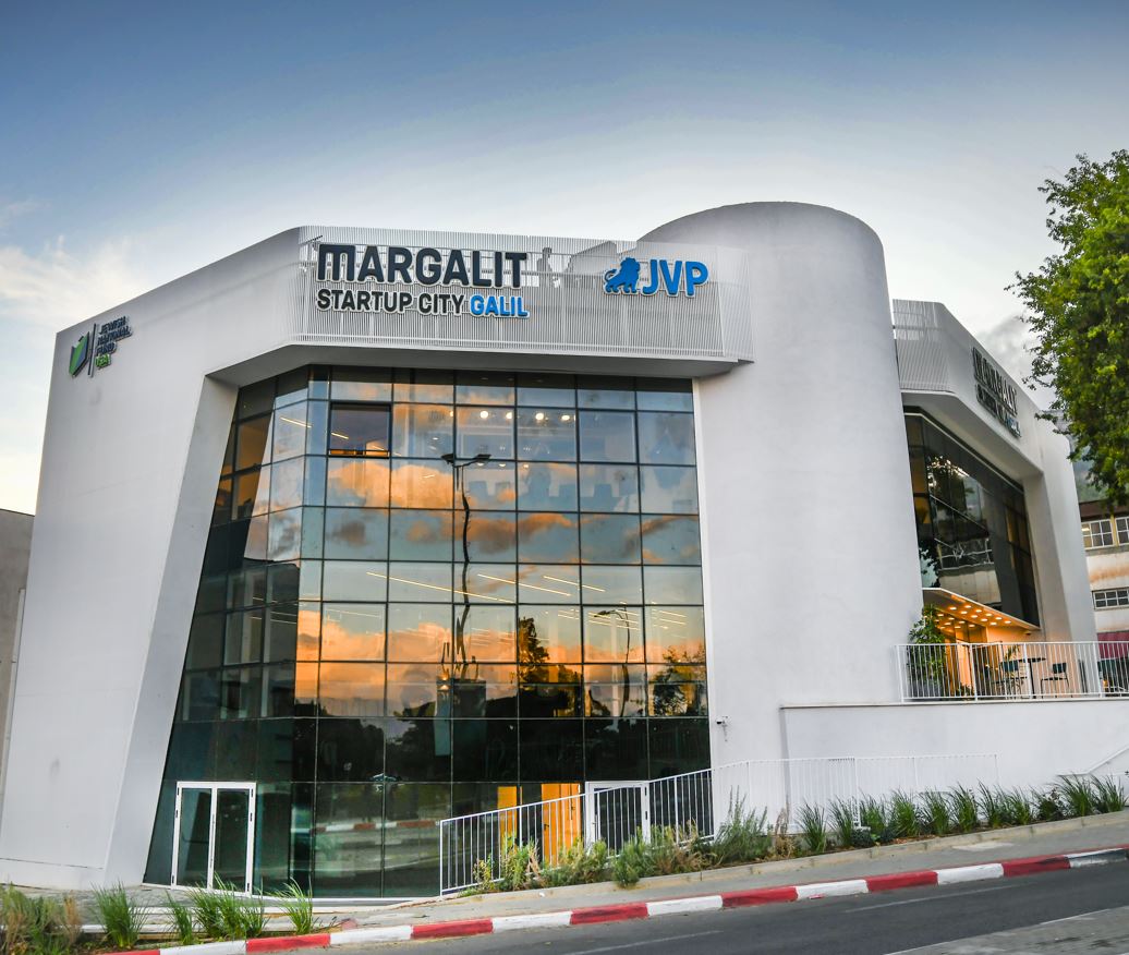 A modern, white building with large glass windows reflects the sky, featuring the signs Margalit Startup City Galil and JVP. A few trees and a street with a red and white curb are visible in the foreground.
