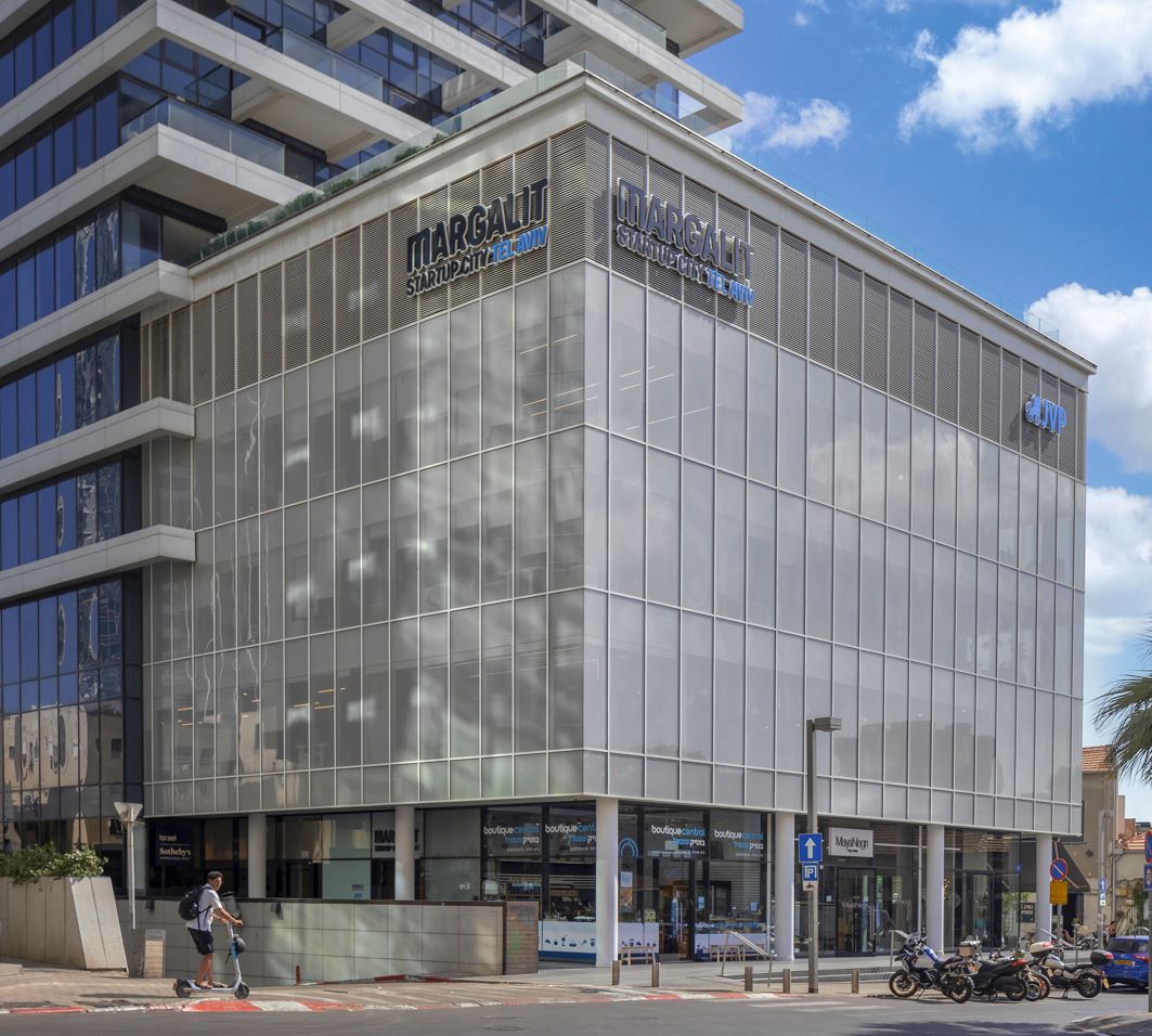 A modern glass building with MARGALIT STARTUP CITY signage. The structure has multiple floors with reflective windows. People and motorcycles are visible at street level. The sky is partly cloudy.