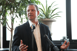 A man in a black suit speaks into a microphone, gesturing energetically. He stands in a room with large windows and a potted plant in the background, discussing Israels dynamic role in venture capital investment.
