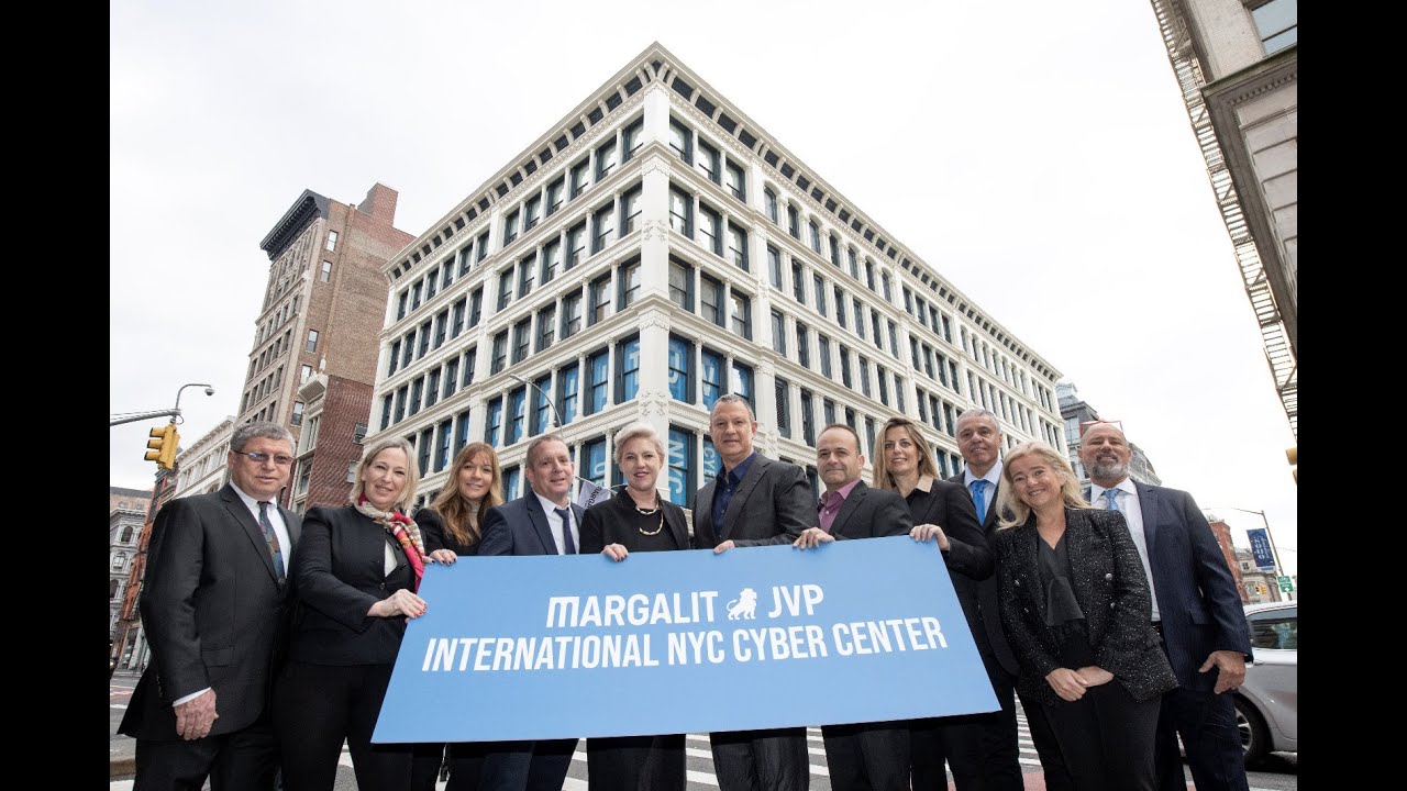A group of people gathers outside a large, windowed building at the bustling street corner, holding a sign that reads Margalit JVP International NYC Cyber Center. This iconic center stands as a testament to innovation and collaboration.