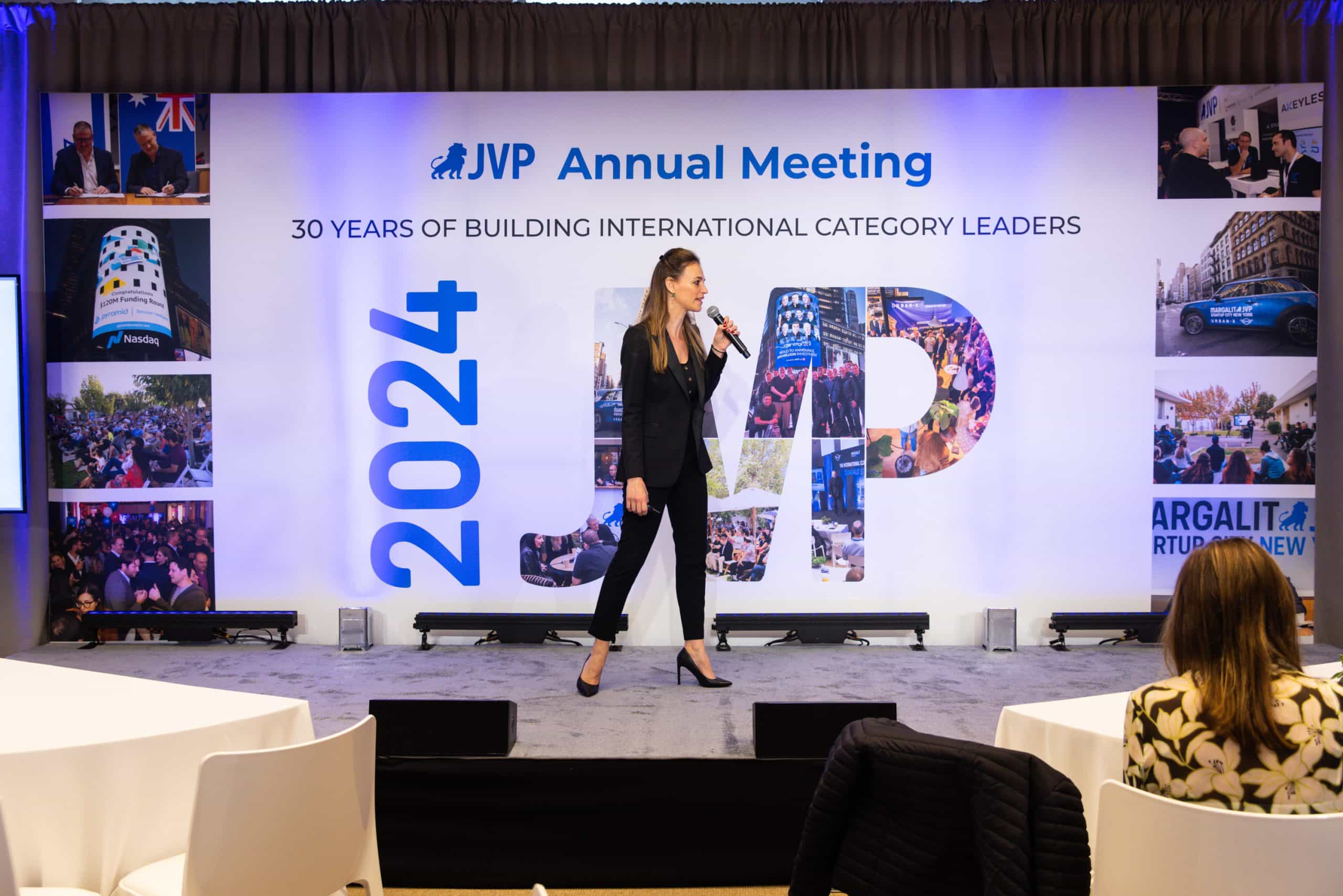 A woman in a black suit speaks on stage at the JVP Annual Meeting 2024, with a large backdrop displaying 30 Years of Building International Category Leaders and event photos.