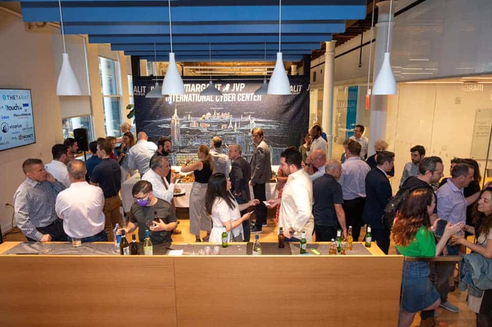 A large group of people socialize and network at an indoor professional event with drinks. A banner in the background reads International Cyber Center. Some people are standing at a counter and talking in small groups.