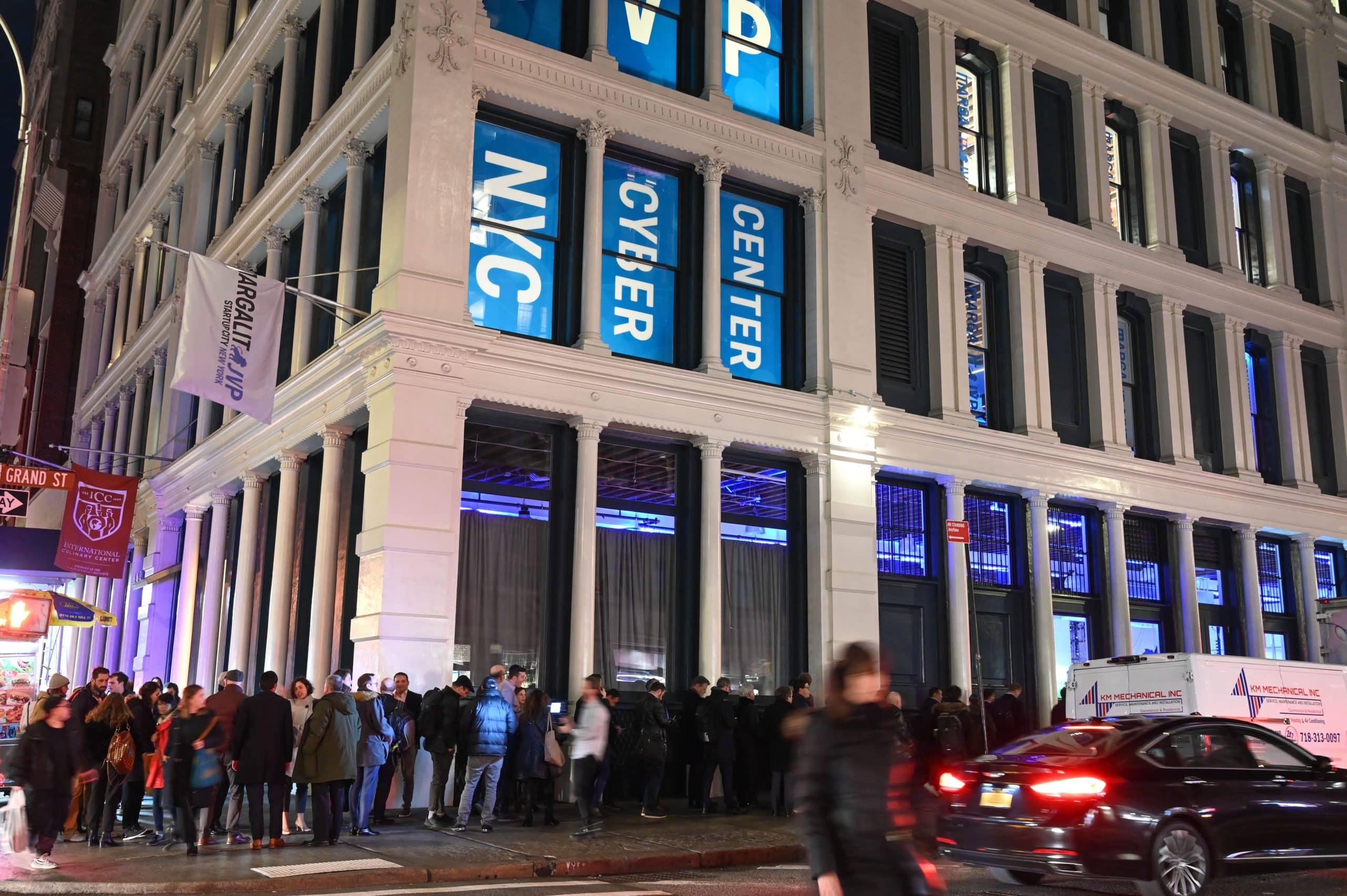 A long line of people waits outside a brightly lit building with large blue “NYC CYBER CENTER” banners at night; cars and pedestrians pass by on the busy street.