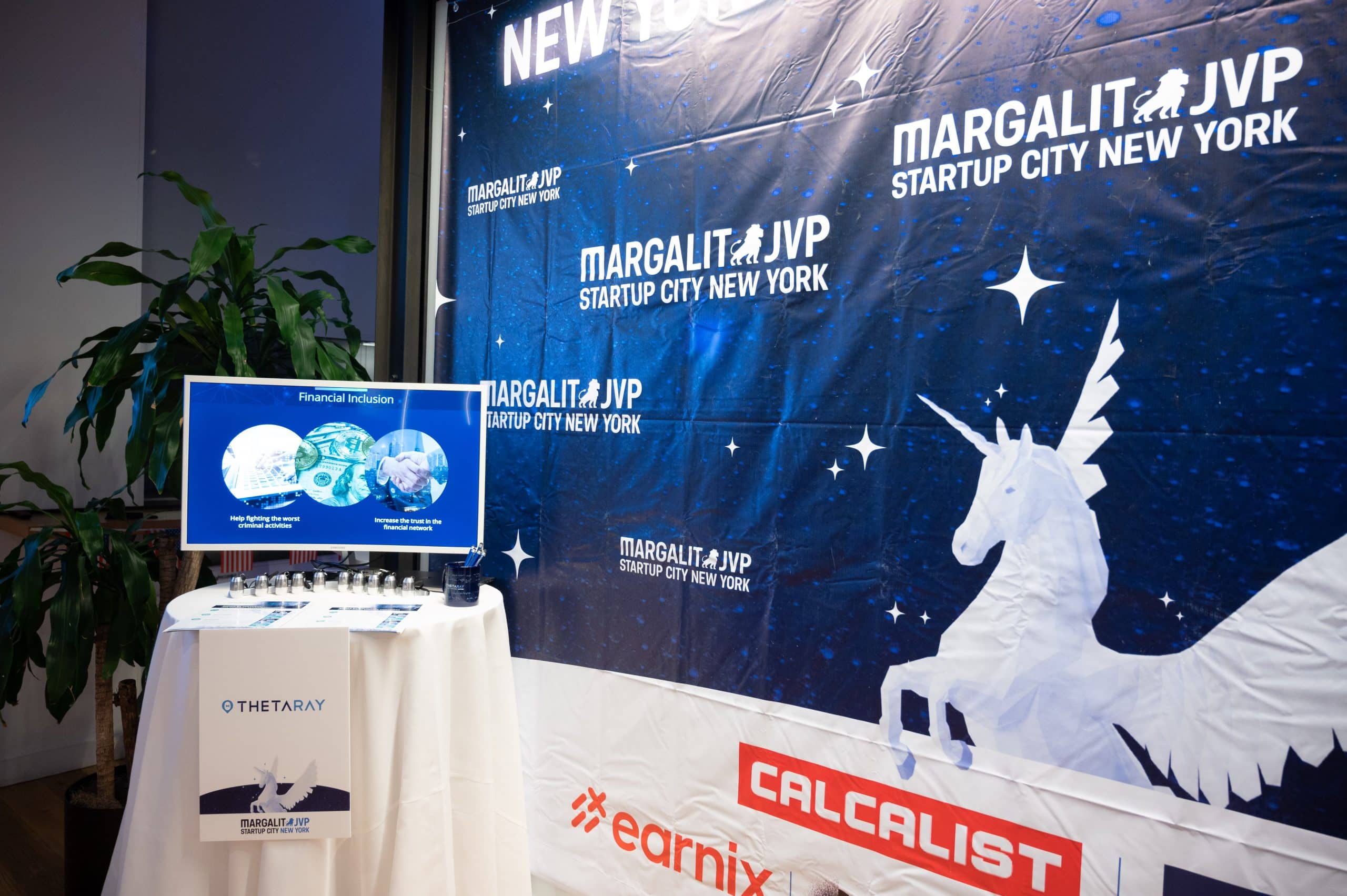A display booth at an event features a blue banner with a white unicorn, Margalit JVP Startup City New York logos, and a screen showing a financial inclusion presentation. Nearby are branded materials and a potted plant.