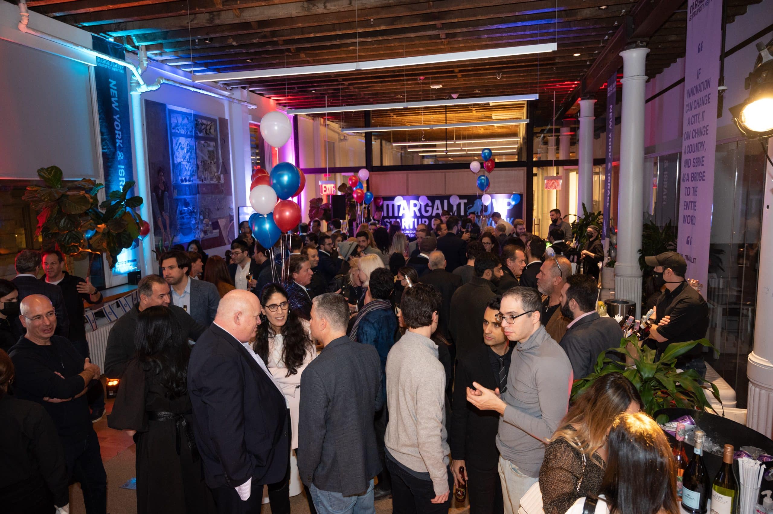A large crowd socializes at an indoor event with colorful balloons and bright lighting. People are mingling, chatting, and some are standing near tables. The space has exposed ceilings and modern decor.