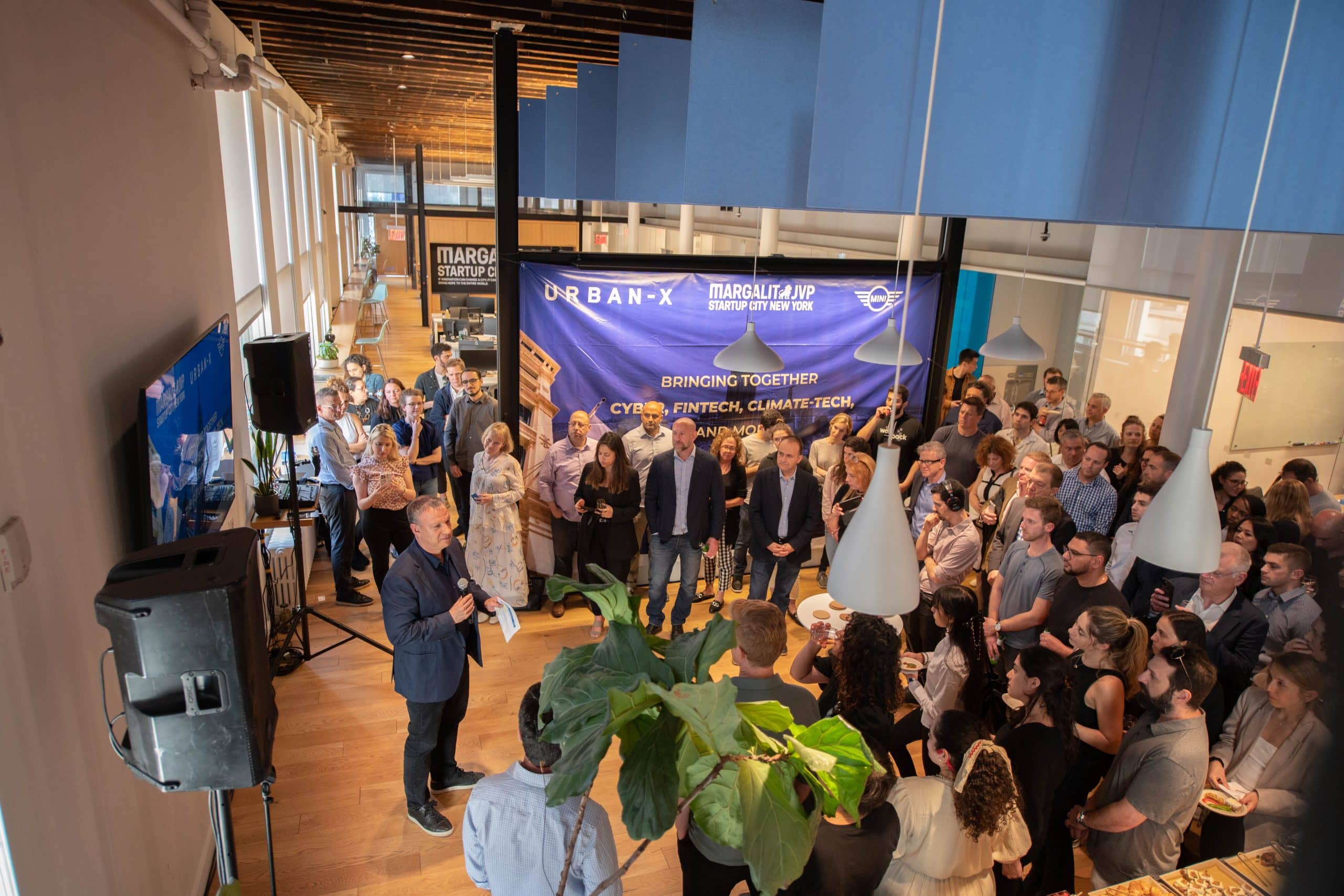 A large group of people gather in a modern, open indoor space with high ceilings, listening to a speaker holding a microphone. A screen and banners about urban and climate tech are visible in the background.