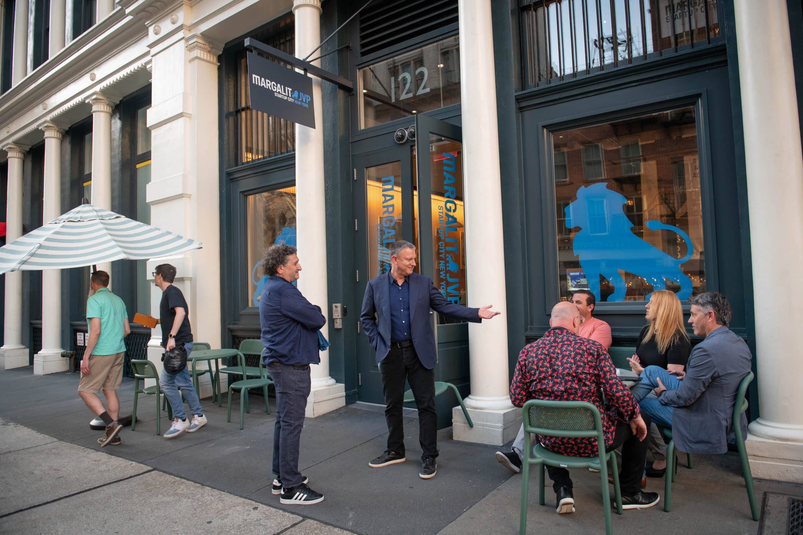 A group of people talk and sit at tables outside a cafe with large windows and blue signage on a city street. Some are standing and gesturing while others are seated and engaged in conversation.