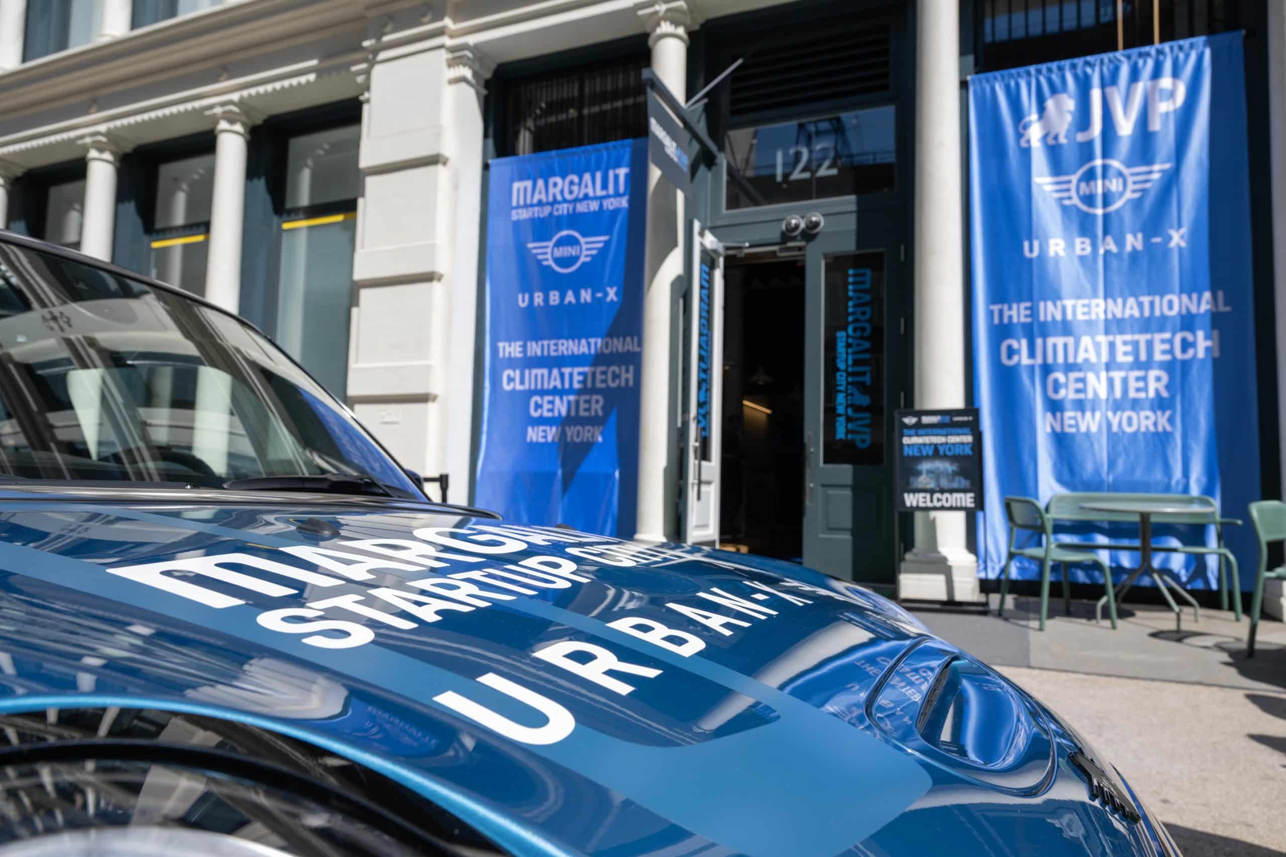 A blue car with “STARTUP URBAN-X” written on the hood is parked outside a building with large blue banners reading “THE INTERNATIONAL CLIMATETECH CENTER NEW YORK” and “MARGALIT STARTUP CITY”.