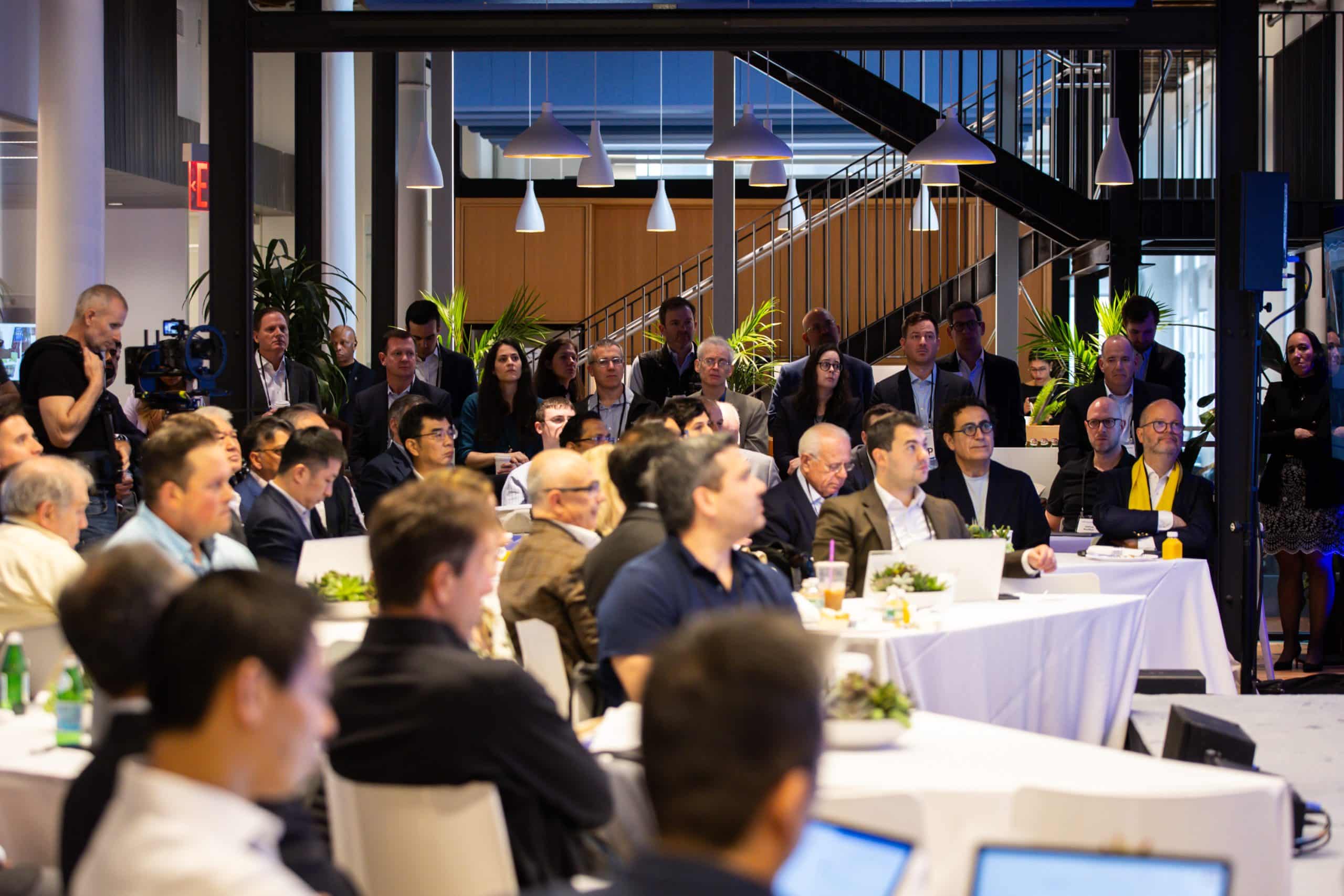A group of people in business attire attend a conference or seminar in a modern, well-lit room. Some are seated at tables with laptops and notepads, while others stand in the background near a staircase.