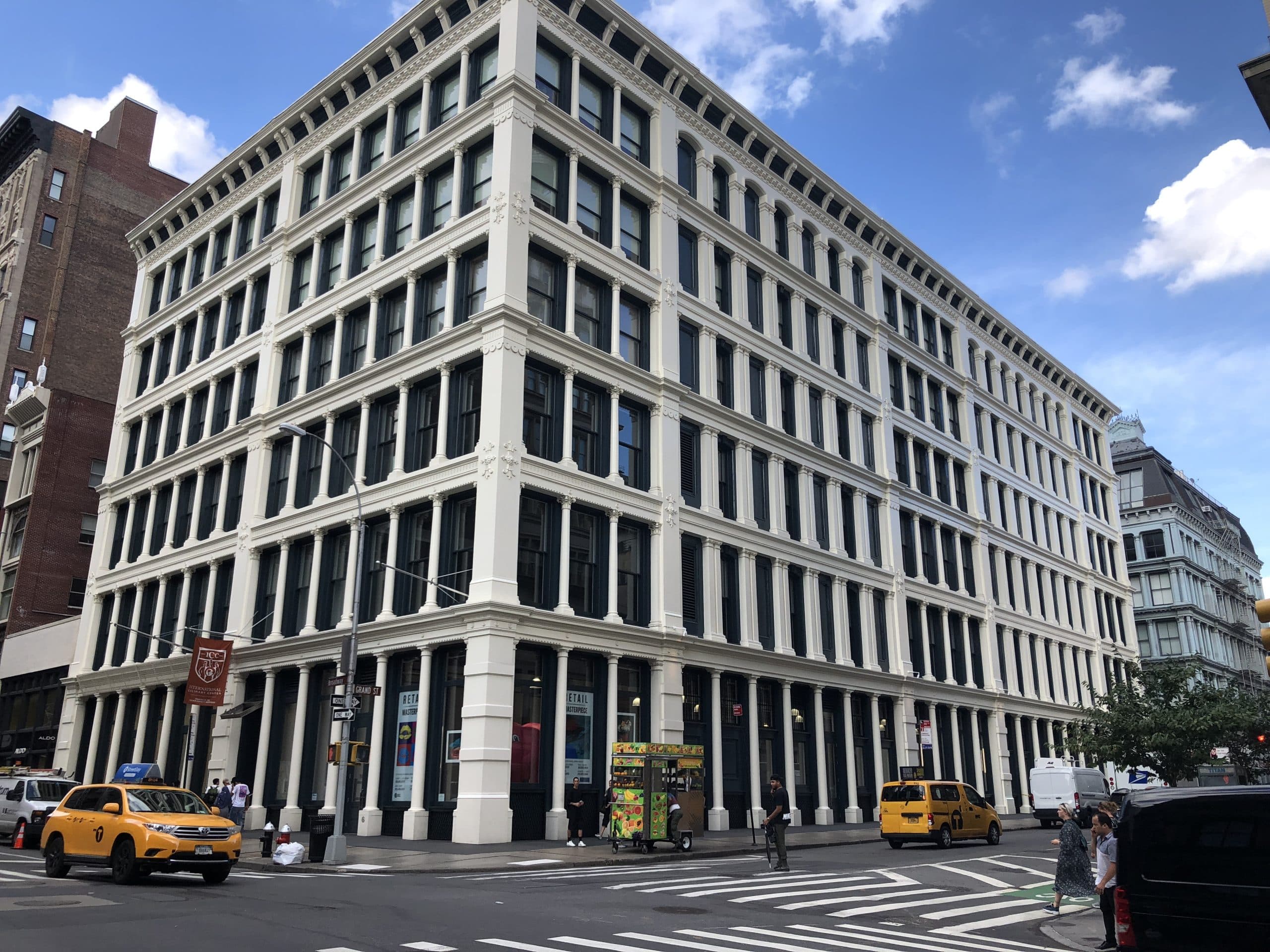 A large white and black historic building with many windows stands at a city street corner in New York. Yellow taxis, a food cart, and pedestrians are visible under a partly cloudy sky.