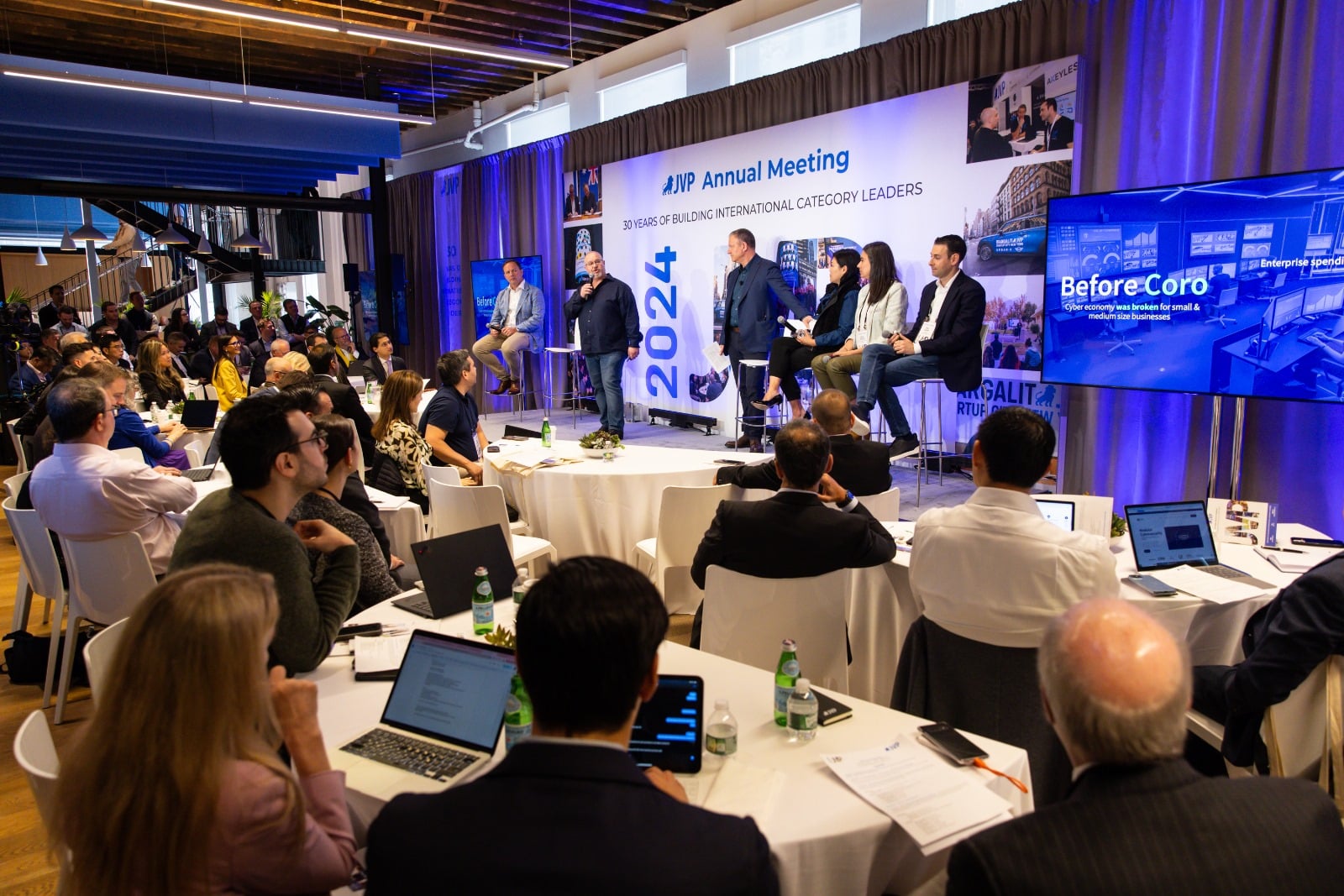 A group of panelists speak onstage at the AIMP Annual Meeting as an audience listens and takes notes at round tables filled with laptops, notebooks, and drinks. Large screens display presentations and signage.