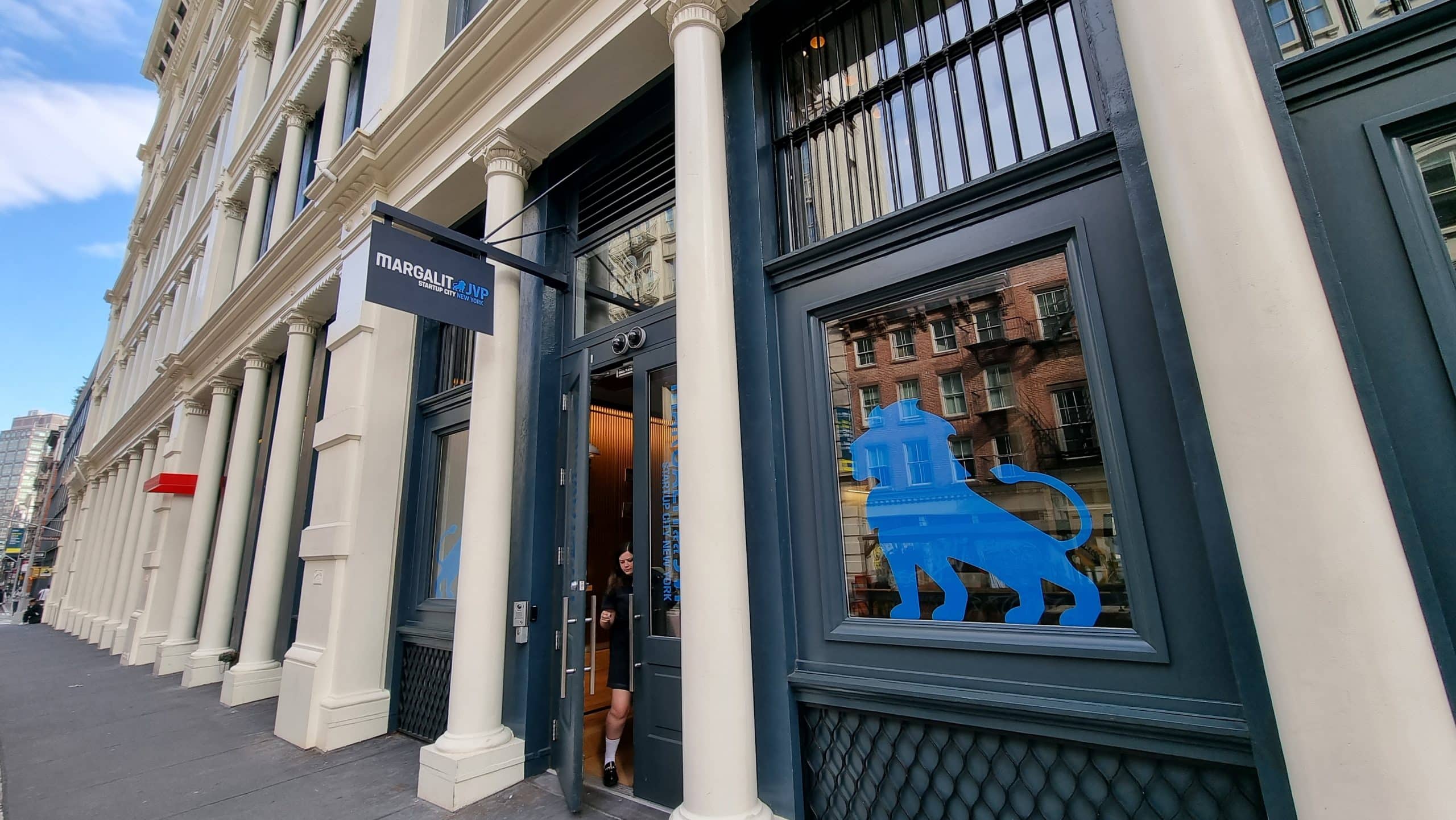 A person exits the ParabolaLAPP storefront with tall cream columns, large windows, and a blue lion graphic on the glass in a city street setting.