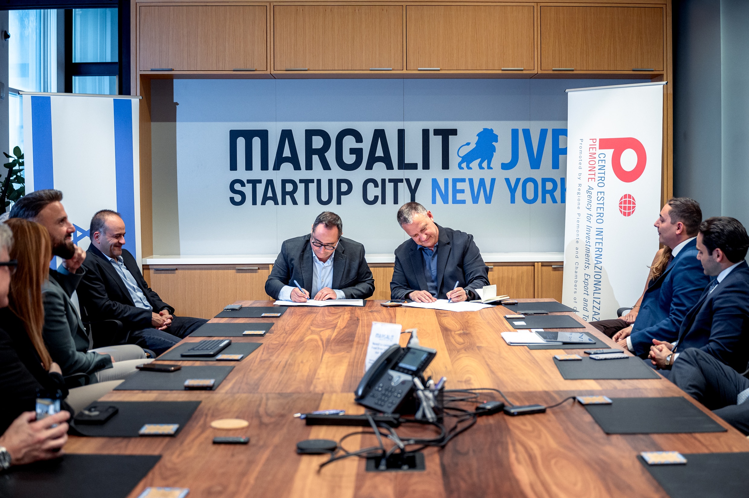 A group of business professionals sit around a conference table as two men in the center sign documents at Margalit Startup City New York, with a banner for the Foreign Press International Organization visible in the background.