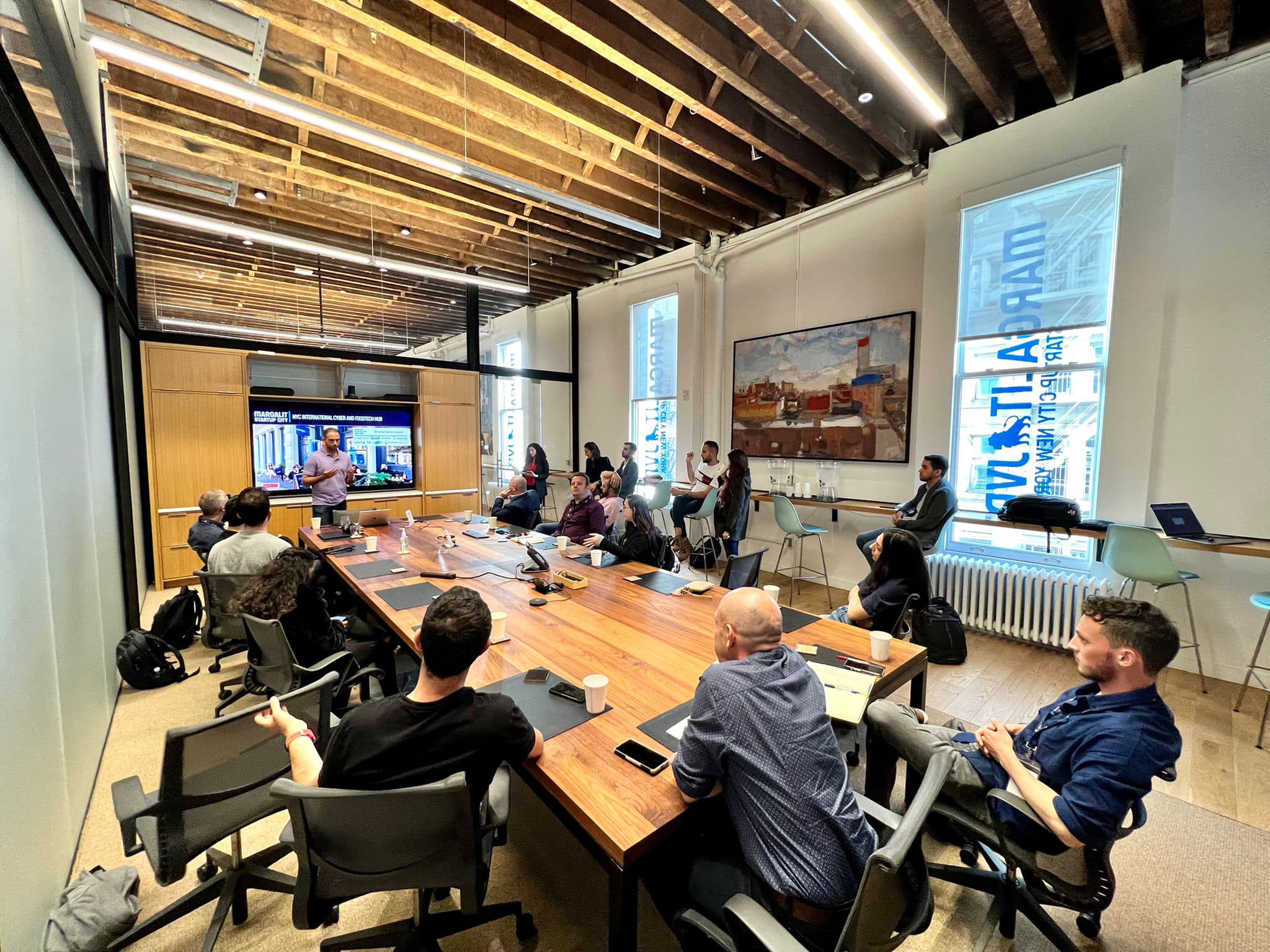 A group of people sit around a long conference table in a modern meeting room, attentively listening to a presenter standing near a screen displaying graphs and charts. The room features exposed beams and large windows.