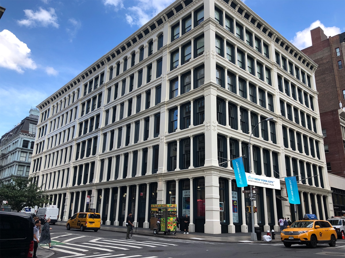 A large, white historic building with tall windows stands on a busy city corner. Yellow taxis, pedestrians, and a food cart are visible on the street under a blue sky with clouds.